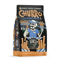 The front of a 12 ounce bag of Bones Coffee Company Churro coffee. Its flavor is churro, and it has a skeleton in front of a churro stand holding out churros on the art.