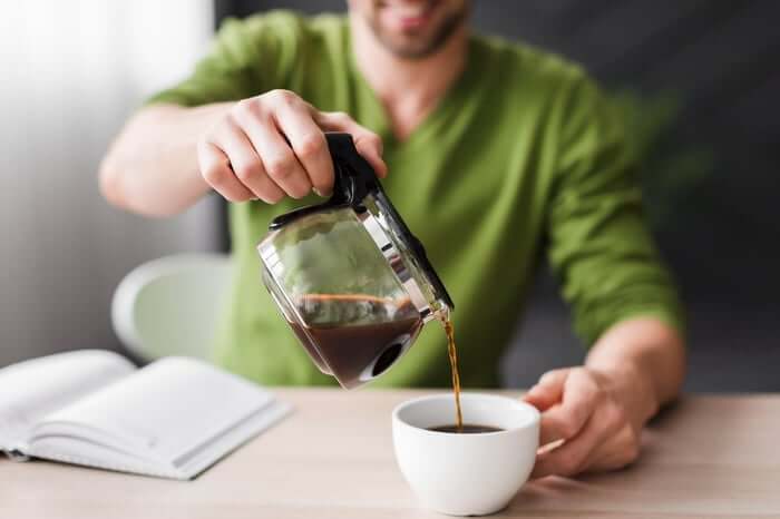 Man pouring hot coffee into cup while reading book on table.