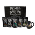 Five four-ounce bags of flavored coffee are in front of a black box that says Bones Coffee company. Beside the bags of coffee is a black hand-thrown mug with the Bones Coffee Company skull logo. The names on the bags are Army of Dark Chocolate, Holy Cannoli, Salty Siren, S’morey Time, and Highland Grog.