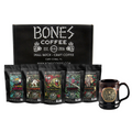 In the background is a black box that says Bones Coffee Company. In front of it are five four ounce bags of coffee and a mug. The coffees are named Guatemala, Ethiopia, Sumatra, Colombia, and Costa Rica. The mug has Bones Coffee’s classic logo on it and is colored black with an orange glaze on top.